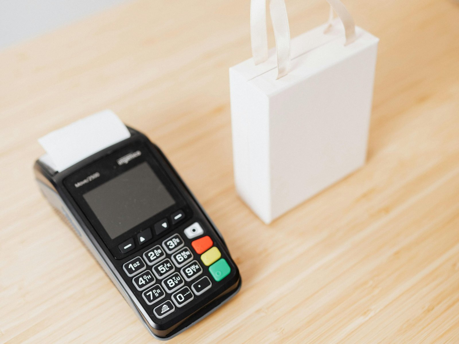 A payment terminal and a small white gift bag on a wooden countertop, ideal for retail or shopping themes.